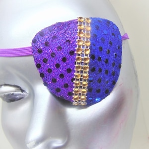 Könnte beinhalten: Ein violettes und blaues Pailletten-Augenpflaster mit einem goldenen Strassband. Das Augenpflaster ist an einem violetten Gummiband befestigt.