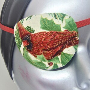 Puede incluir: Un parche para el ojo de tela con un diseño de pájaro cardenal rojo y hojas de acebo verdes sobre un fondo blanco. El parche para el ojo tiene una correa elástica.