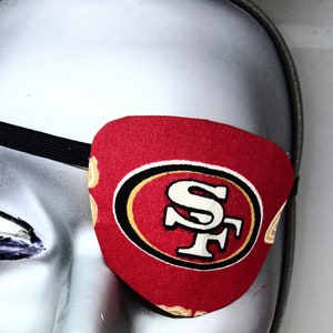 May include: A red eye patch with a gold and black San Francisco 49ers logo. The eye patch is worn on a mannequin with a silver face and red lips.