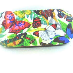 Puede incluir: Una funda rígida para gafas blanca con un estampado de mariposas de colores. La funda está decorada con mariposas de varios colores, incluyendo rojo, azul, verde, amarillo y morado.