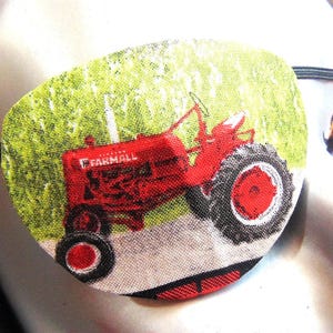 May include: A red and black Farmall tractor eye patch with a green field background. The tractor is driving through a field of green grass.