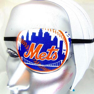 Może przedstawiać: Biały łatanka na oko z niebiesko-pomarańczowym logo New York Mets. Logo przedstawia piłkę baseballową z nazwą drużyny i panoramą miasta.