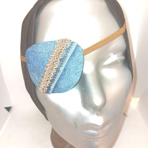 May include: A blue denim eye patch with gold trim, attached to a brown elastic headband.