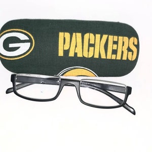 以下が含まれることがあります： 緑と黄色の布製の眼鏡ケースに「PACKERS」の文字が印刷されています。黒いフレームの老眼鏡がケースの上に置かれています。