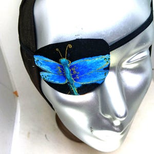 May include: A black fabric eye patch featuring a hand-painted bright blue dragonfly with gold-colored outlines and antennae. The patch is secured with a black elastic strap and is displayed on a silver-colored mannequin head form.