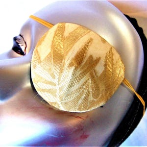May include: A gold eye patch with a floral pattern. The eye patch is made of fabric and has an elastic strap.