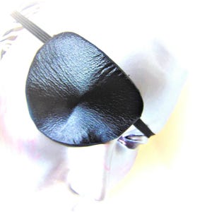 Könnte beinhalten: Schwarze Augenklappe aus Leder mit einem Gummiband. Die Augenklappe ist oval und hat eine strukturierte Oberfläche. Das Gummiband ist schwarz und verläuft um den Kopf.