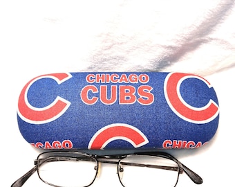 Puede incluir: Un estuche azul para gafas con las palabras "CHICAGO CUBS" en letras rojas y blancas. El estuche tiene un patrón repetido de la letra "C" en rojo con un contorno blanco. Un par de gafas con montura negra descansa frente al estuche.