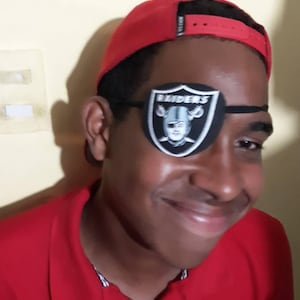 Peut inclure: Une jeune personne portant une chemise rouge et un cache-œil Raiders noir et blanc. La personne porte également une casquette de baseball rouge avec une visière noire.