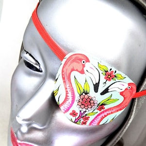 May include: A pink and green eye patch with a flamingo print. The eye patch is made of fabric and has an elastic strap.