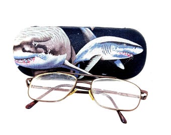 May include: Eyeglasses with a brown metal frame and clear lenses rest in front of a shark-themed glasses case. The case is black with illustrations of two sharks in shades of gray and blue.