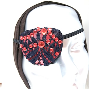 May include: A decorative eye patch featuring a navy blue lace design with red accents and embellishments. The eye patch is adorned with red rhinestones and is attached to a black elastic band for wear.