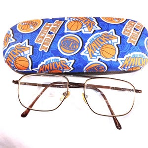 May include: A pair of brown metal eyeglasses with square lenses and a blue and orange patterned hard case with the words "New York Knicks" repeated on the case.