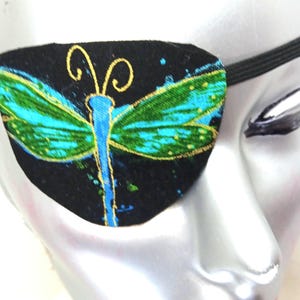 May include: A black eye patch with a colorful dragonfly design. The dragonfly has green wings with gold accents and a blue body.