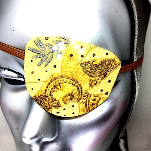 May include: A yellow eye patch with a brown paisley pattern and silver accents. The eye patch is made of fabric and has an elastic band.