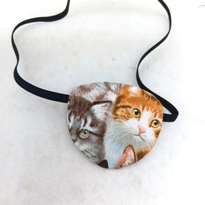 Könnte beinhalten: Eine Augenklappe mit einem Stoffdruck von zwei Katzenköpfen, einer grauen und einer orange-weißen, die mit einem schwarzen Gummiband verbunden sind. Die Augenklappe ist zum Abdecken eines Auges bestimmt.