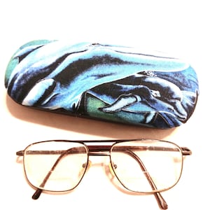 May include: A blue and black fabric eyeglasses case with a whale design. The case is closed and sitting on a white surface. A pair of brown metal eyeglasses with clear lenses is also pictured.