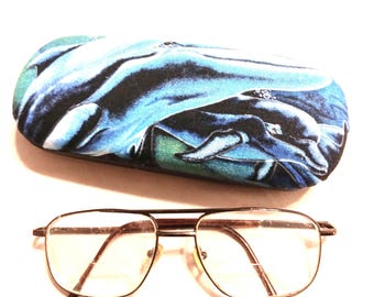 May include: A blue and black fabric eyeglasses case with a whale design. The case is closed and sitting on a white surface. A pair of brown metal eyeglasses with clear lenses is also pictured.