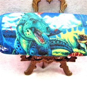 Peut inclure: Un étui à lunettes en tissu bleu et vert à thème dinosaure avec un dinosaure vert à la bouche rouge et aux yeux jaunes. L'étui est sur un support en métal doré.