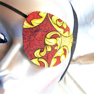 May include: A red and gold eye patch with a floral pattern. The eye patch is made of fabric and has an elastic strap.