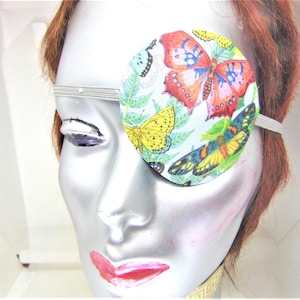May include: A decorative eye patch featuring a colorful butterfly design. The round patch is adorned with various butterflies in shades of red, yellow, and blue, set against a light blue background with green foliage. It is held in place with a white elastic band.