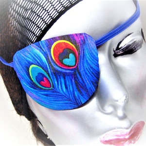 May include: A blue eye patch with a peacock feather design. The patch has a bright blue background with colorful peacock feathers and a pink heart in the center of each feather.