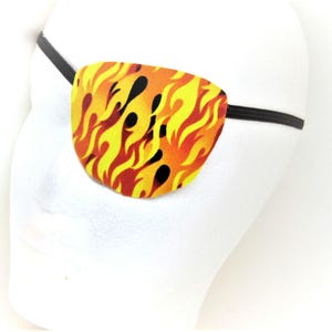 Puede incluir: Un parche en el ojo con un diseño de llamas en amarillo, naranja y negro. El parche se sujeta con una banda elástica negra. El parche está sobre una cabeza de maniquí blanca.