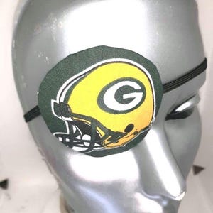May include: Green Bay Packers eye patch featuring a green and yellow football helmet design. The eye patch is made of fabric and has an elastic strap.