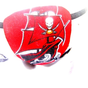 Puede incluir: Parche de ojo de pirata rojo con un diseño de barco y calavera en blanco y negro. El barco tiene una vela roja y una calavera dorada con espadas cruzadas.