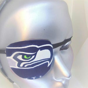 May include: A blue and white eye patch with a green eye and a Seattle Seahawks logo. The eye patch is worn on a mannequin's head.