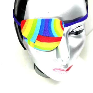 May include: A colorful rainbow eye patch with a blue elastic strap. The eye patch is made of fabric and has a geometric pattern.