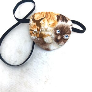 May include: An eye patch featuring a printed fabric design of three cats, including a tabby, a ginger cat, and a Siamese cat with blue eyes. The patch is oval-shaped and attached to a thin, black elastic strap for securing around the head.