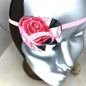 May include: An eye patch featuring a pink and red rose floral pattern with a pink awareness ribbon overlay. The patch is secured by a thin pink elastic strap that wraps around a dark brown headband worn on a silver-colored mannequin head.