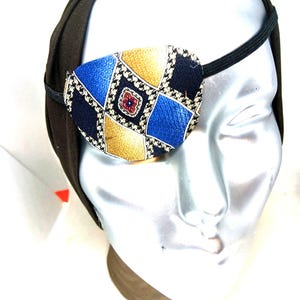 May include: An eye patch with a geometric pattern in blue, gold, and black, attached to a black elastic band. The eye patch is worn on a mannequin head with a dark green head covering.