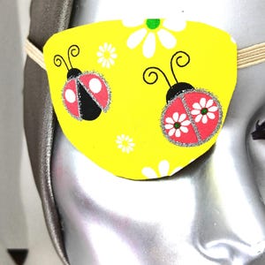 May include: A yellow eye patch with two ladybugs and white flowers. The ladybugs have black spots and red wings.