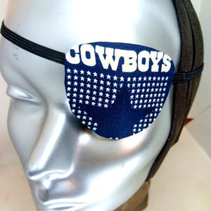 May include: A navy blue eye patch with the word "COWBOYS" in white block letters. The patch is decorated with white stars and is attached to a black elastic band. The eye patch is designed to be worn over one eye.