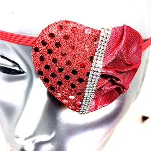 May include: A red eye patch with sequins and rhinestones. The patch is shaped like a heart and has a ruffled edge.