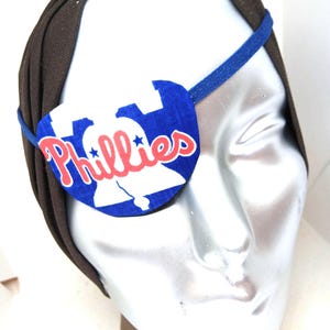 May include: Blue and white eye patch with the word "Phillies" in red lettering and a white star on each side of the word. The eye patch is worn on a mannequin's head.