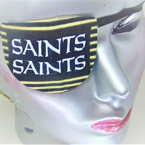 Puede incluir: Parche para el ojo a rayas negras y doradas con el texto "Saints Saints" impreso en blanco.