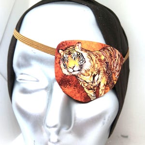 May include: A brown eye patch with an orange and yellow tiger print. The eye patch is worn on a mannequin's head.