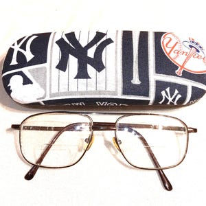 May include: A brown metal eyeglasses frame with clear lenses. The eyeglasses are sitting on a white surface in front of a black and white fabric eyeglasses case with a New York Yankees logo and baseball theme.