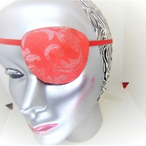 May include: A red eye patch with a floral pattern, worn over a mannequin's eye. The eye patch is held in place with a red elastic band.