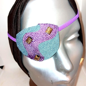 May include: Purple and blue eye patch with gold accents and an elastic strap. The eye patch is worn on a mannequin head.