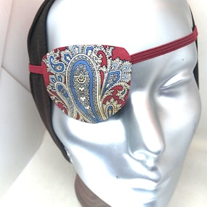 May include: A decorative eye patch with a paisley pattern in red, blue, and cream colors. The eye patch is attached to a maroon elastic band and a brown headband. The eye patch is on a mannequin head.