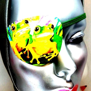 May include: A green eye patch with a colorful image of three frogs on a green background. The frogs are yellow, green, and brown with black eyes.