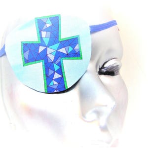 May include: A blue eye patch with a green outline and a mosaic cross design. The eye patch is made of fabric and has an elastic strap.