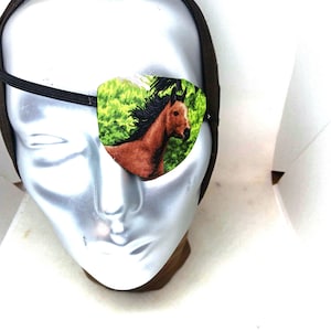 Puede incluir: Un parche ocular de tela con un caballo marrón de crin negra sobre un fondo de follaje verde brillante, sujeto con una banda elástica negra y colocado sobre una cabeza de maniquí plateada con una diadema de color marrón oscuro.