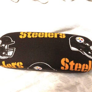 以下が含まれることがあります： 黒いメガネケース。黄色いステンシルフォントで「Steelers」と「Steel」の文字。ケースには、白黒のフットボールヘルメットの画像と、スティーラーズのロゴが描かれています。
