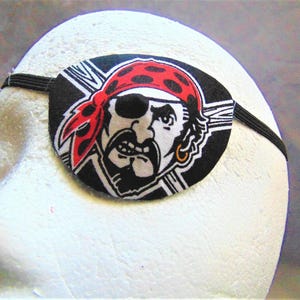 May include: Black eye patch with a pirate illustration. The pirate is wearing a red bandana with white polka dots and has a gold earring. The eye patch has an elastic strap.