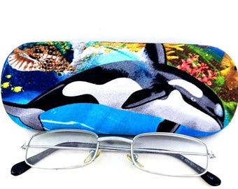 May include: A blue and white glasses case with a colorful image of an orca whale swimming in the ocean. The case is made of soft fabric and has a zipper closure. A pair of silver-rimmed rectangular glasses are sitting on top of the case.